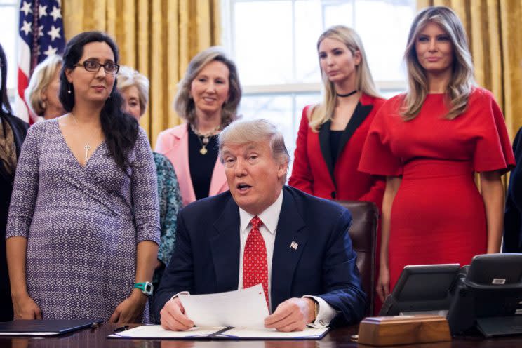 Trump holds papers at his desk with red-dress-wearing Melania, among others, standing behind him.