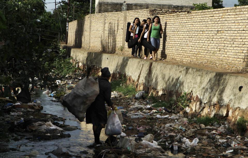 In this Dec. 11, 2012 photo, young women carry their instruments along the edge of a polluted stream where a woman scavenges for recyclable goods in the garbage, as they head to their practice session with "The Orchestra of Instruments Recycled From Cateura," in Cateura, a vast landfill outside Paraguay's capital of Asuncion, Paraguay. The community of Cateura could not be more marginalized. But the music coming from garbage has some families believing in a different future for their children. (AP Photo Jorge Saenz)