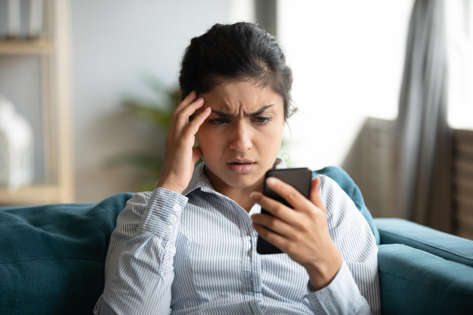 Confused millennial Indian girl sit at home look at cellphone having operational problems, frustrated young woman feel stressed with slow Internet connection, virus attack or spam on smartphone