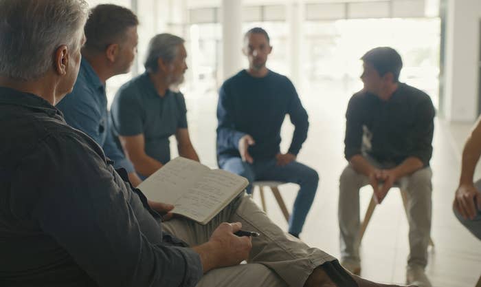 A group of six people in casual attire sits in a circle during a discussion meeting. One person holds a notebook.