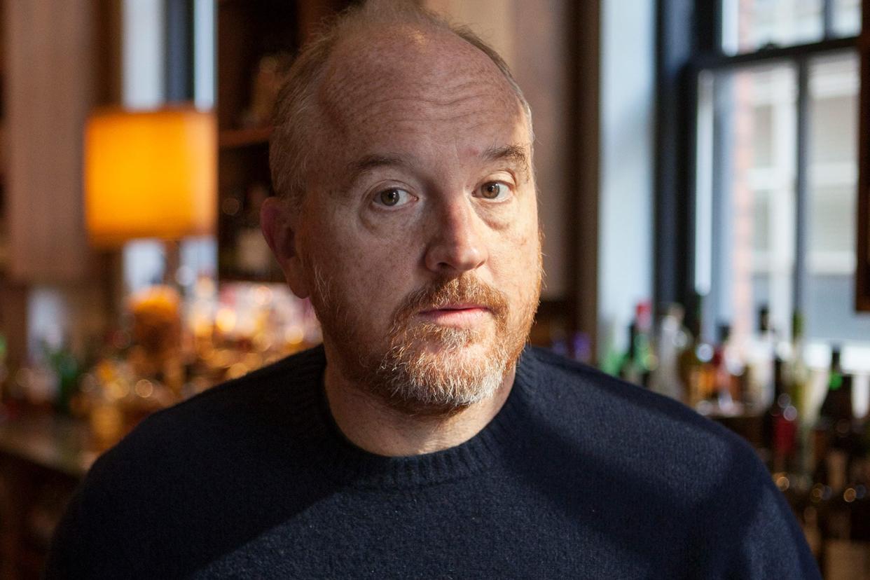 The comedian sits in a bar wearing a dark crew-neck sweater, looking right at the camera.