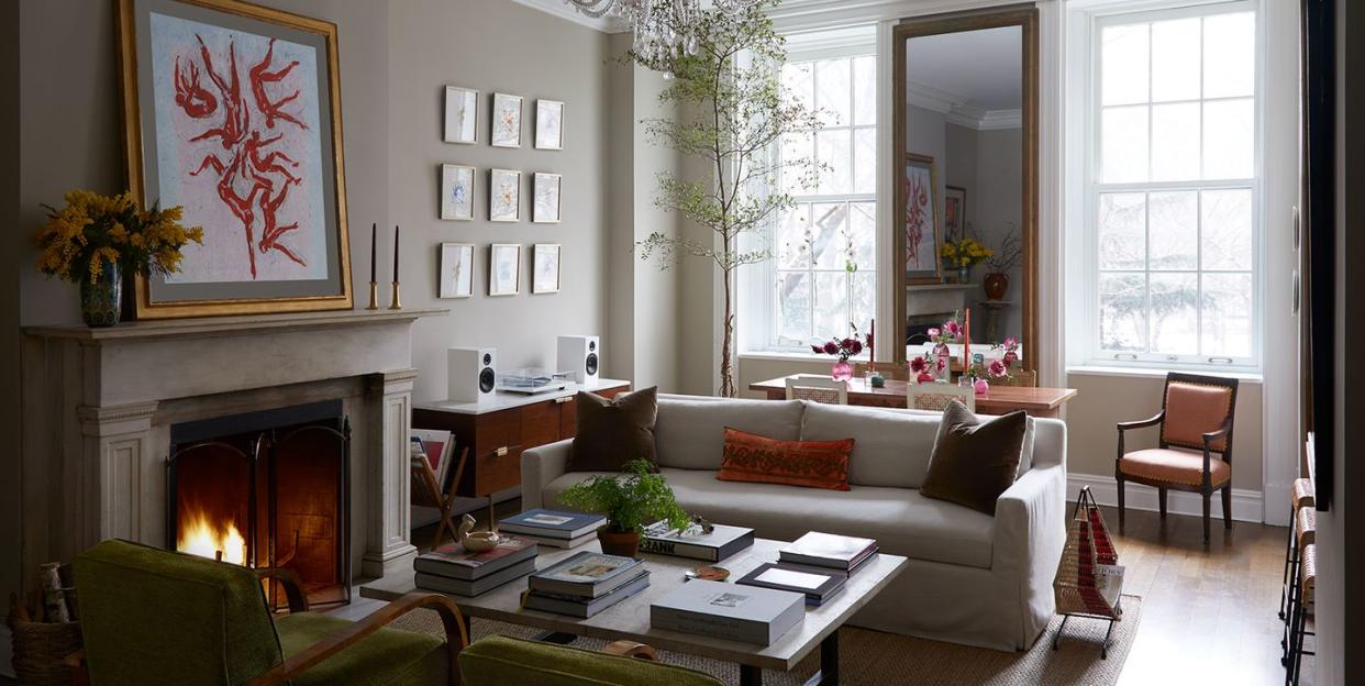 full living view with a seating area with a whitish sofa and two low armchairs opposite and a table at center with books and a lit fireplace with artwork over it on the left and a dining area behind the sofa and tall window and a mirror