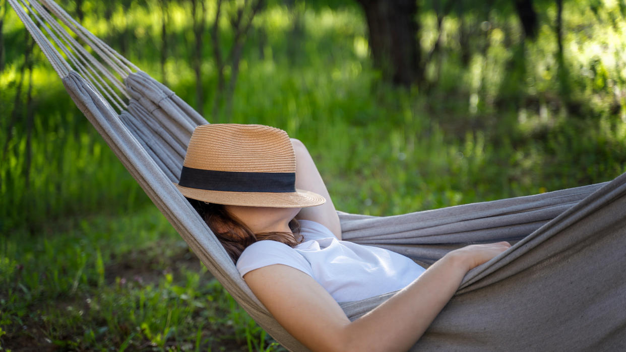  Woman resting in a hammock in a summer garden covering her face with a straw hat. 