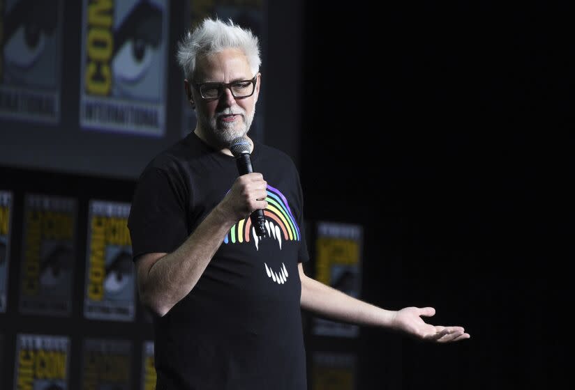 A man with white hair and a beard wearing glasses and a rainbow shirt while speaking into a microphone on a stage