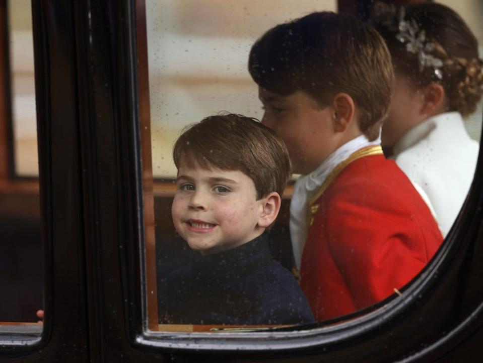 Prince Louis, Prince George, and Princess Charlotte of Wales depart the coronation service at Westminster Abbey in a carriage. Louis smiles at a camera.