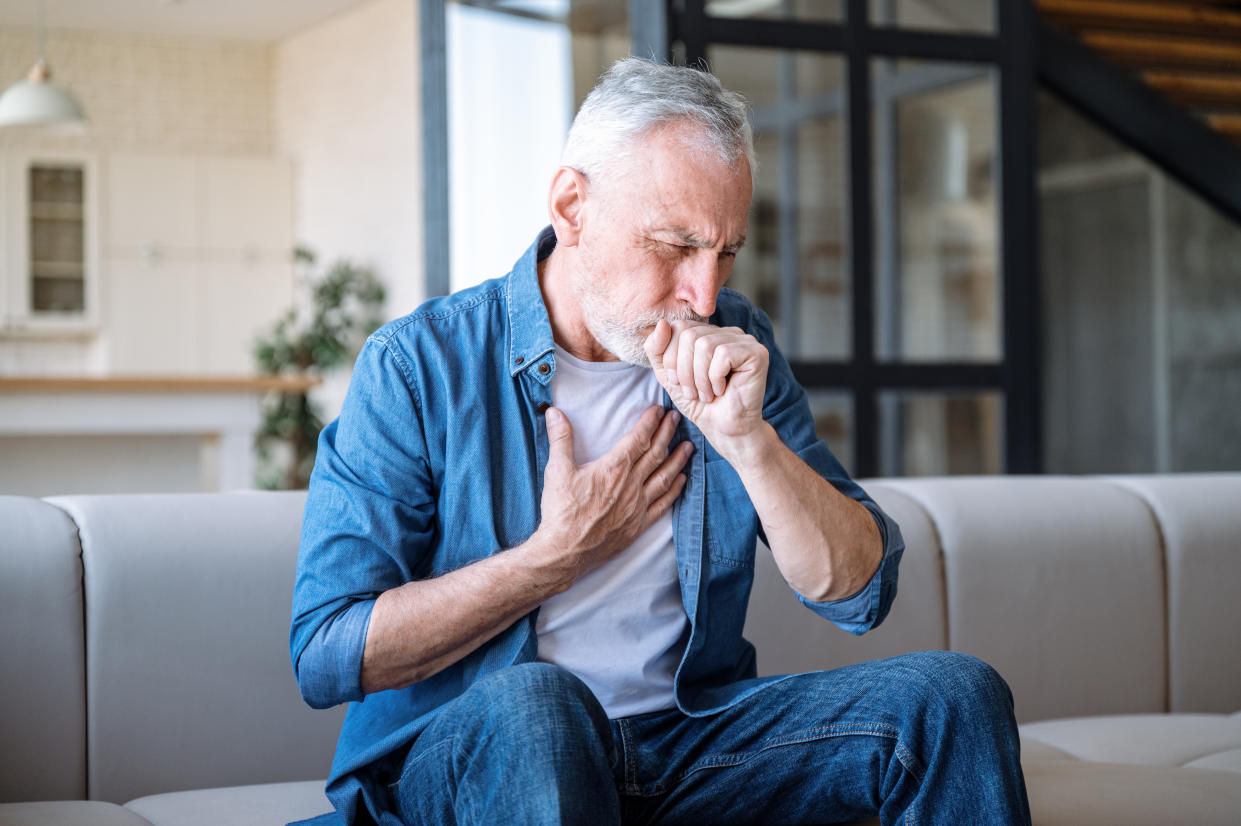 Senior man sitting on sofa at home and holding hand on chest. Male having asthma attack, difficulties with breathe, feeling severe pain or dyspnea�