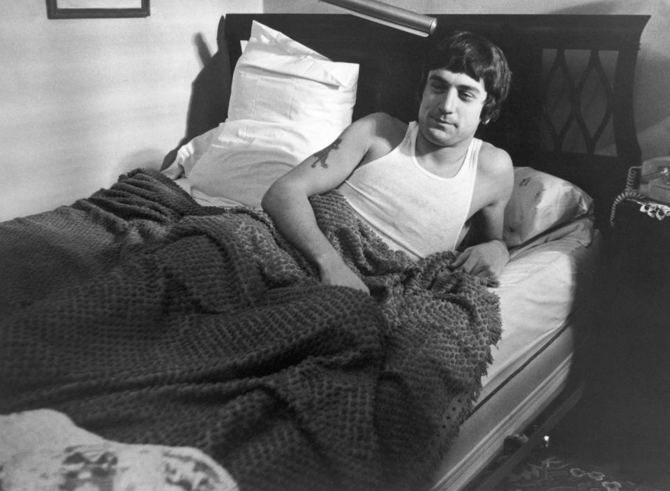 robert de niro in character for mean streets lies in bed on one elbow, he wears a white tank top and has a blanket over his body to his waist