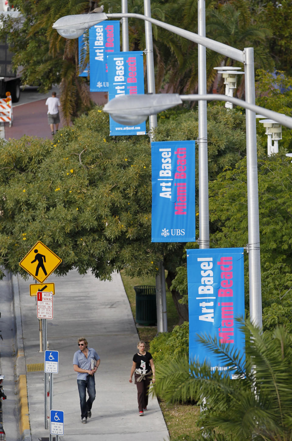 Pedestrians walk under banners announcing Art Basel Miami Beach, Tuesday, Dec. 4, 2012 in Miami Beach, Fla. Art Basel Miami Beach and about two dozen other independent art fairs open Thursday. Tens of thousands of people are expected through Sunday at the fairs throughout Miami and South Beach. (AP Photo/Wilfredo Lee)