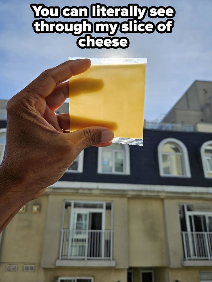 A hand holds a slice of cheese in clear packaging against a backdrop of a multi-story building with arched windows