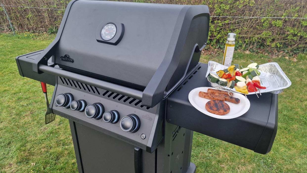  An example of how we test grills, The Napoleon Phantom Rogue on test, with a burger, some sausages, and vegetable kebabs resting on its side table. 