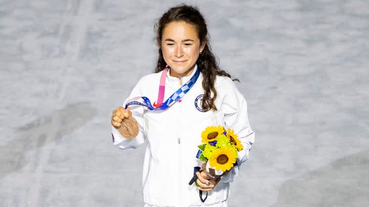 A runner receives a bronze medal a the Olympics wearing a white track suit and holding yellow flowers