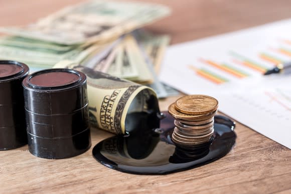 Wooden surface on which are a pair of small oil barrels, a stack of bills, a small pool of black liquid, a stack of pennies, and a piece of paper with graphs.