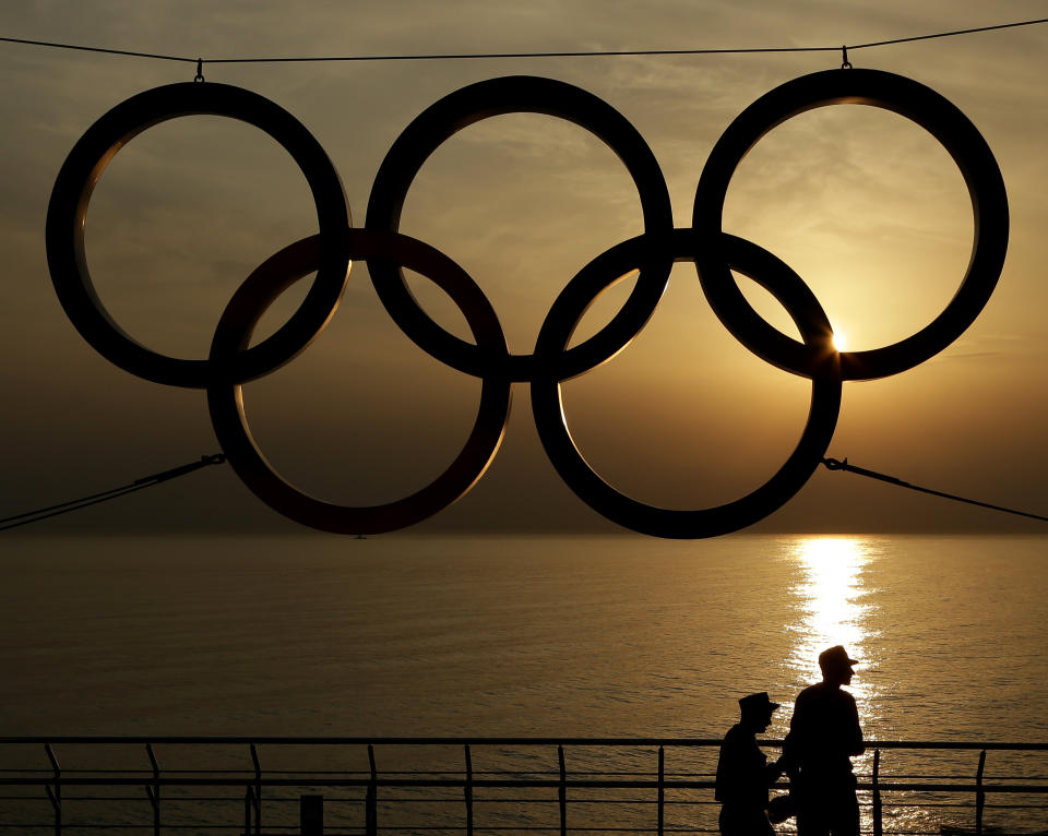 FILE - In this Monday, Feb. 24, 2014 file photo, People stand under a set of Olympic Rings as they watch the sun set over the Black Sea a day after the close of the 2014 Winter Olympics, in Sochi, Russia. U.S. athletes trying to make the Winter Olympics will have to be fully vaccinated for COVID-19 under a groundbreaking new policy announced Wednesday, Sept. 22, 2021 by the U.S. Olympic and Paralympic Committee.(AP Photo/Charlie Riedel, File)