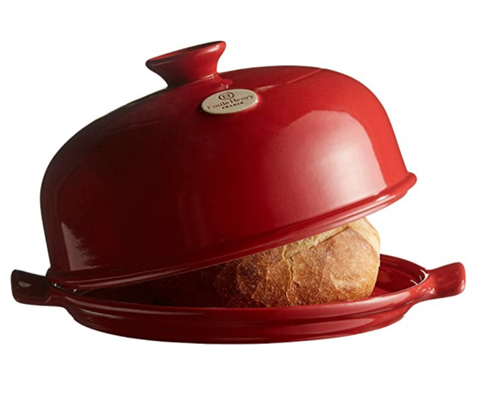 A red Emile Henry clay bread oven sits on a white background with the dome lid slightly open on the right showing a glimpse of a bread loaf.