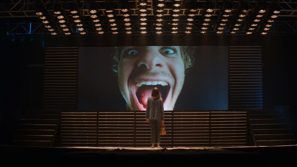 Maya Hawke's Frankie stands in front of a giant projection of Andrew Garfield's Link's face in a still from Mainstream.