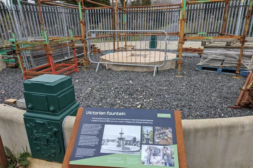 The site of the Victorian Fountain at Belfast City Cemetery with a large empty space surrounded by fencing and scaffolding but with a 'Victorian Fountain' tourism info sign in foreground