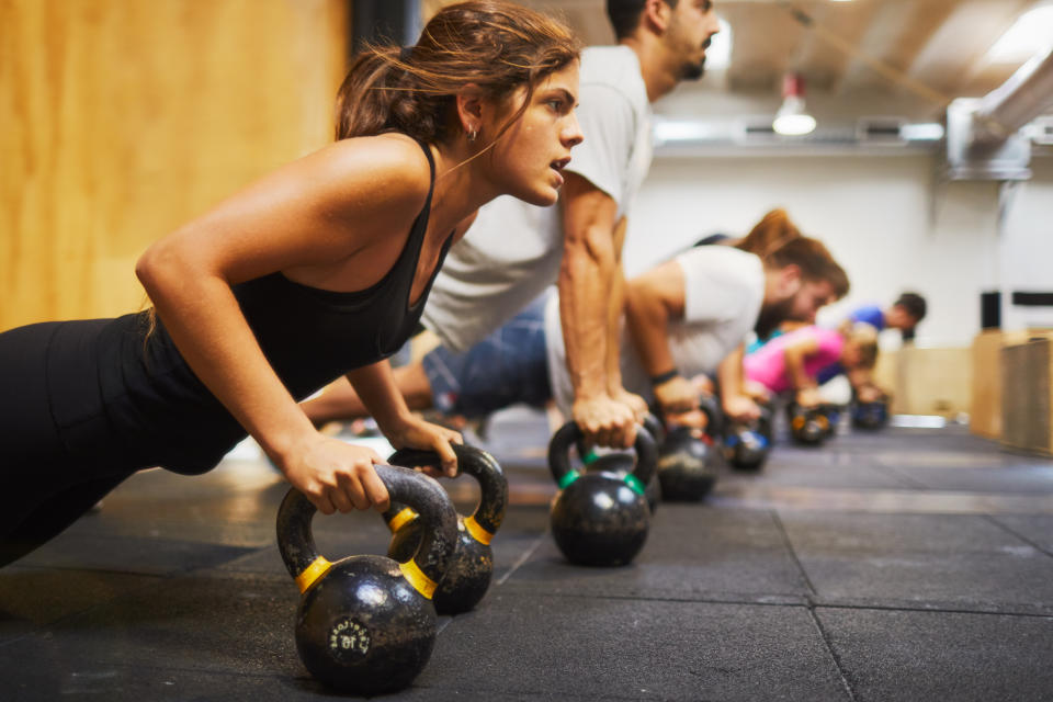 Weight training gym photo of a class using weights and body weight to train. A line of women and men do pushups on kettle bells.