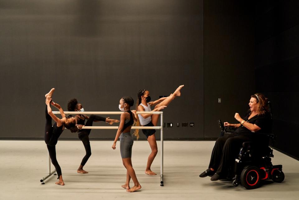 Children rehearse at a ballet barre while an instructor speaks to them.