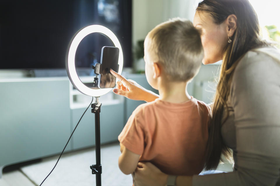 A woman and a young child use a smartphone mounted on a ring light stand, both looking at the phone's screen together