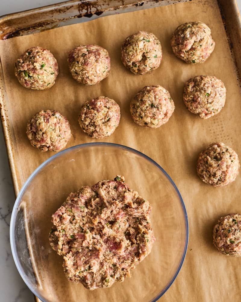 Meatballs on a parchment paper ready for baking, with a bowl of ground beef in a mixing bowl