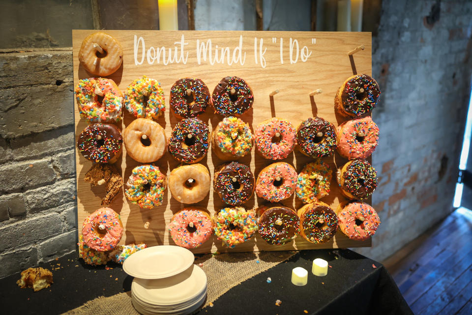 Donut bar display at a wedding with a sign saying "Donut Mind If 'I Do'" and various donuts on pegs. Stacked plates and lit candles nearby