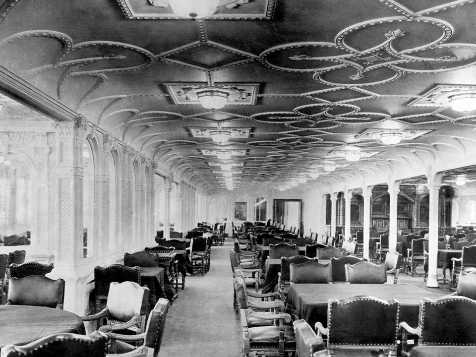 A black-and-white photo of the first-class dining room aboard the Titanic, which is a large room that has large tables surrounded by plush chairs.