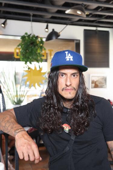 Actor Richard Cabral poses at a coffee shop. He wears a black shirt, has long black hair, and has a Dodgers hat on.