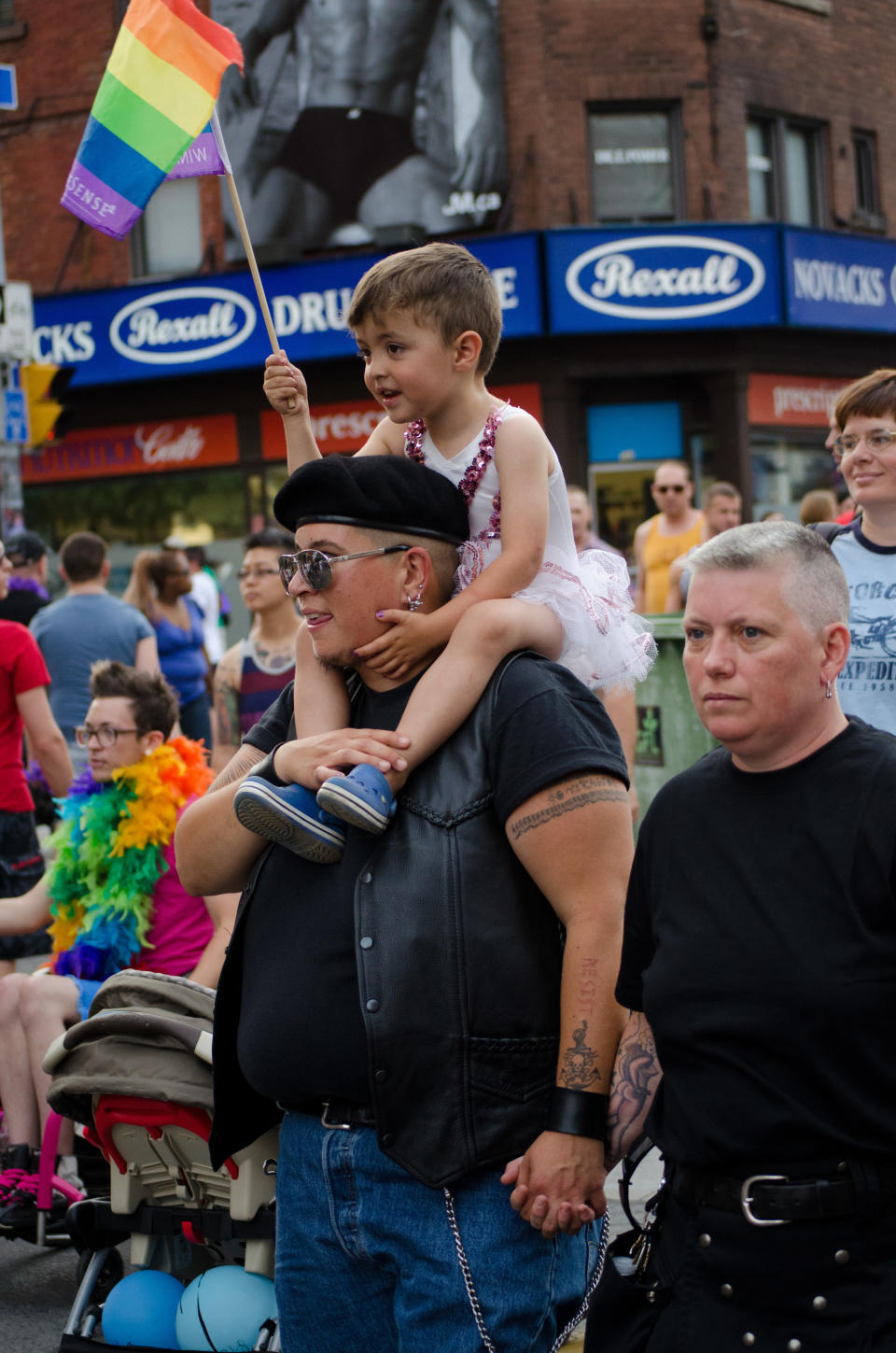 A couple with child on shoulder at a 2012 Pride parade in Toronto, child is wearing a white tutu and waving a rainbow flag against a backdrop of building, signage and people. (Photo by Roberto Machado Noa/LightRocket via Getty Images)