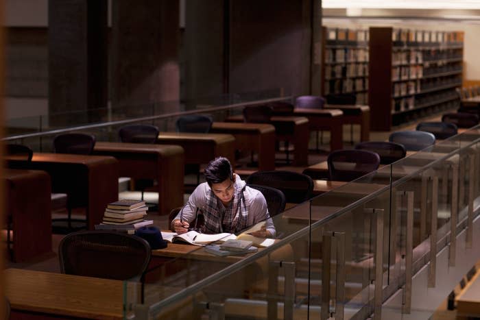 Person sitting alone at a desk in a library, reading a book with several books stacked nearby. The library appears empty and quiet