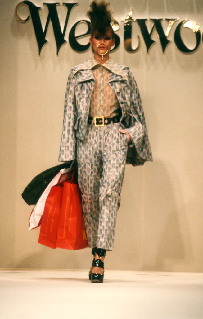 <p>British designer Vivienne Westwood is known to be experimental, creative, and eccentric when it comes to her runway collections. Some may remember the 1993 Spring Summer show in Paris when Kate walked down the runway topless eating a Magnum ice cream bar. She is no stranger to the madness after modeling for Westwood for decades. Here, the model is pictured on the catwalk during the 1994-1995 Autumn Winter fashion show in Paris, France, rocking a patterned co-ord set, gold nose accessory, poofed hair, large belt, shiny black heels, and shopping bags.</p>
