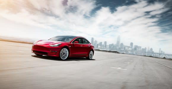 A Tesla Model 3 sedan driving in an empty parking lot. A city skyline is in background.