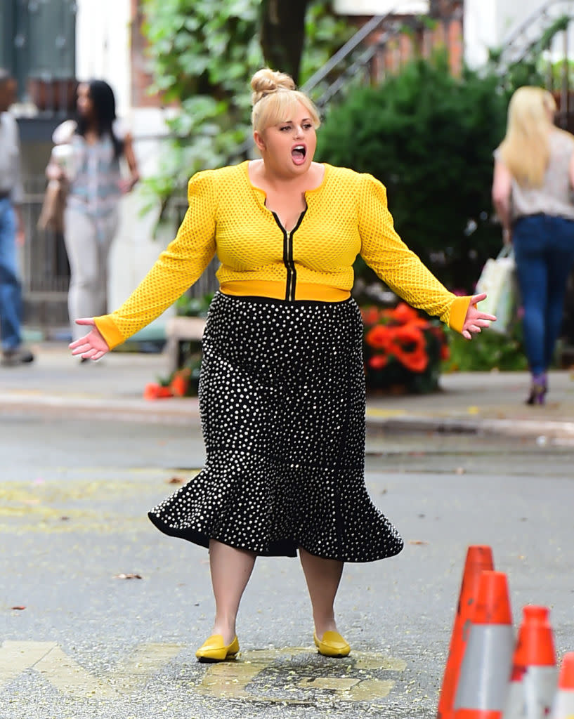 Rebel Wilson on the set of Isn't It Romantic wearing a yellow long-sleeved shirt and sparkly black skirt. 