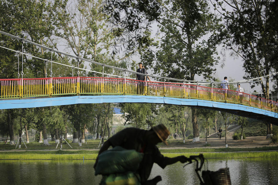 <p>North Koreans walk cross the Pothong River via a suspended bridge while another man is silhouetted as he plants grass on a sidewalk on June 20, 2017, in Pyongyang, North Korea. (Photo: Wong Maye-E/AP) </p>