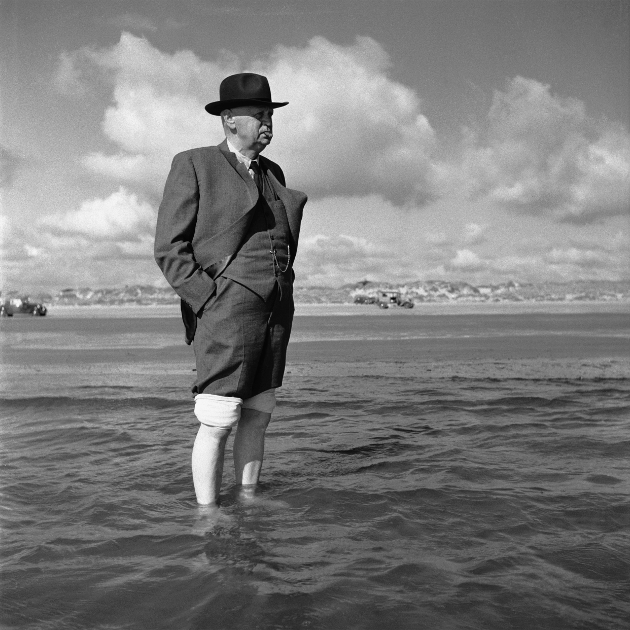 A man rolls up the legs of his suit, paddles into the water to turn his back on the world and stare out to sea