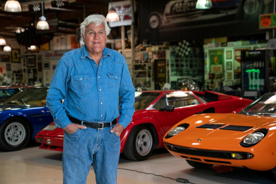 Jay Leno standing in a denim shirt and jeans in front of his exotic car collection