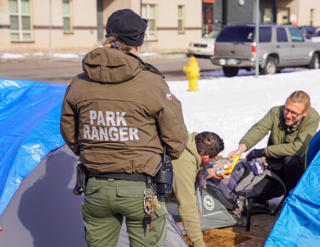 Denver park ranger Jodie Marozas watches as Tom Kaiser, a mental health clinician with WellPower, speaks with an unhoused man living in a tent near a city park in below-freezing weather.
