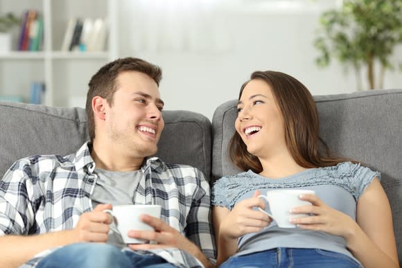 A smiling young couple relaxes on the couch with two cups of coffee.