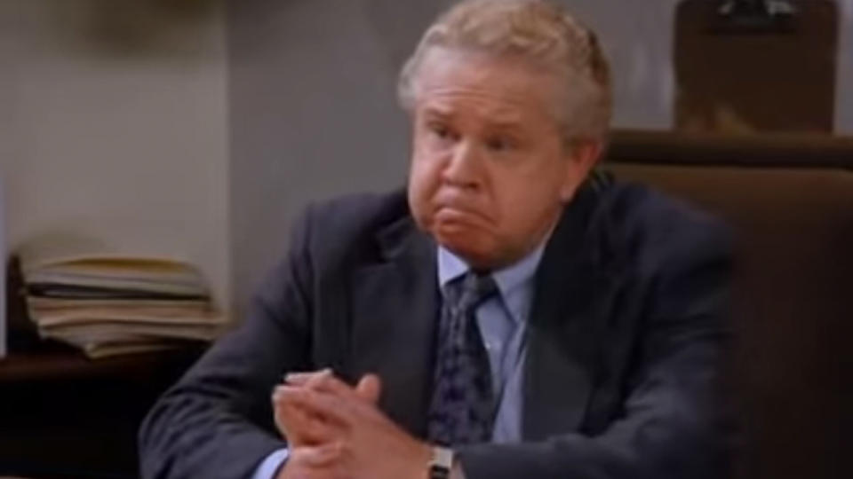 Patrick Cronin sitting at desk, wearing a blue suit in Seinfeld.