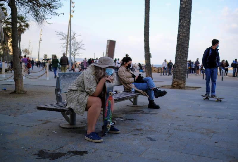 People wearing protective face masks sit on a bench in front of the Barceloneta beach, in Barcelona