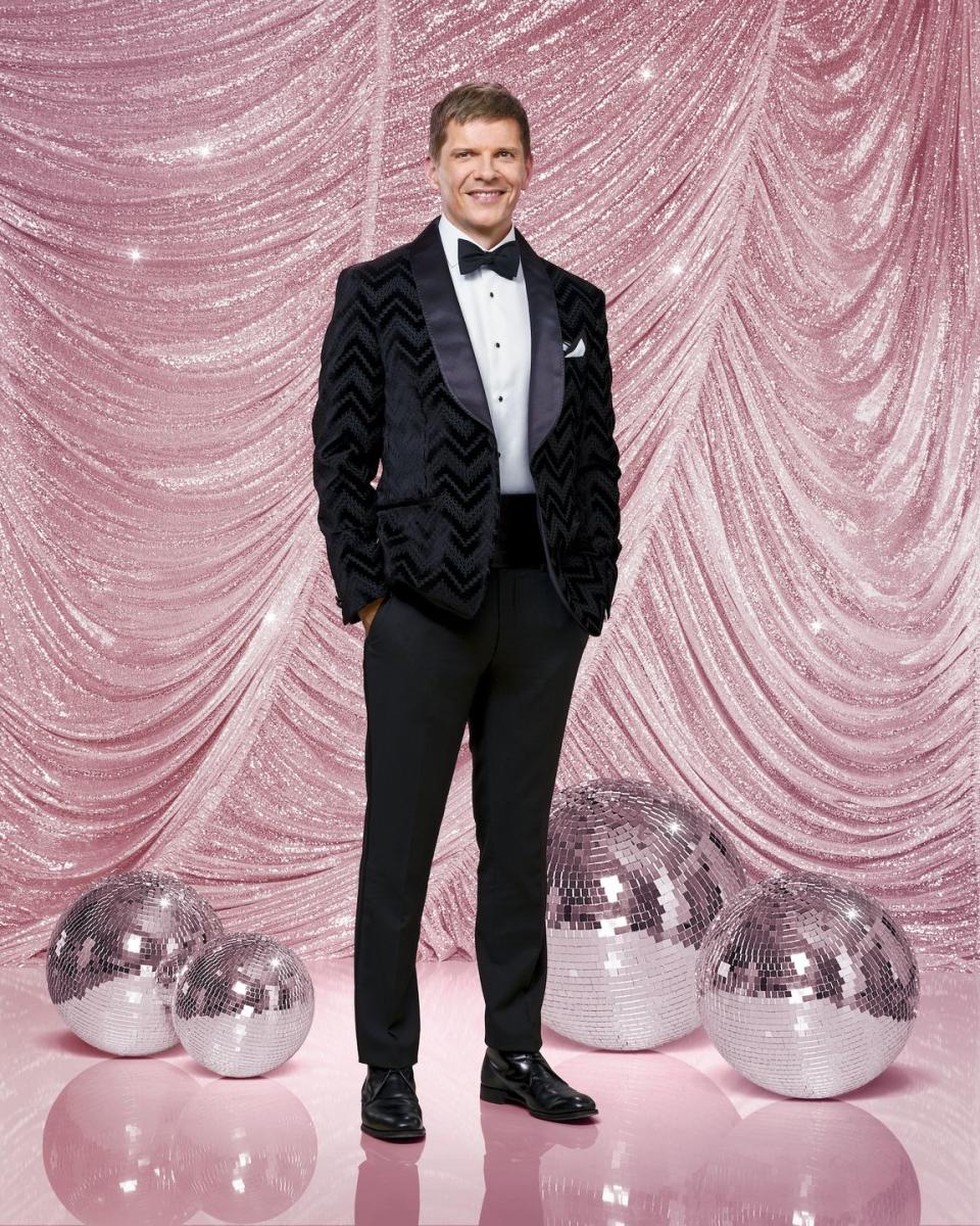 nigel harman for strictly come dancing 2023, a man stands with hands in pockets looking at the camera and smiling, he wears a black suit with bow tie and is surrounded by glitterballs