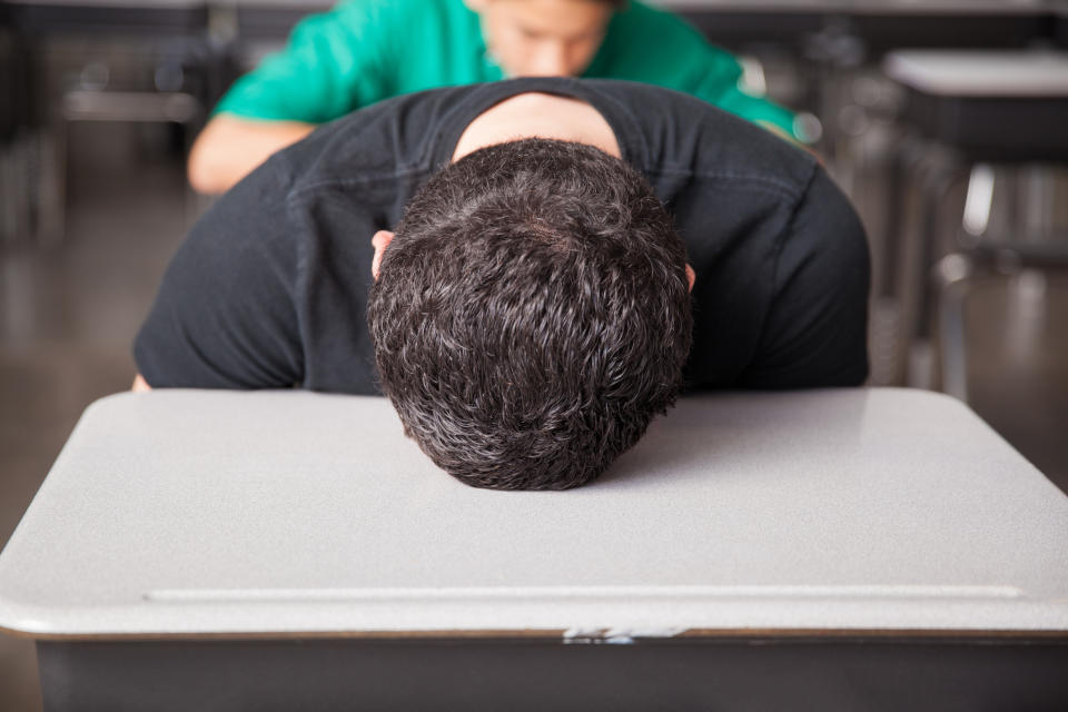 A person with short dark hair leans forward with their head resting on a desk, while another person is seen in the background