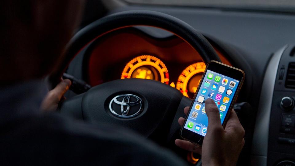 Ein Mann sitzt in einem Auto am Steuer und hält ein Smartphone in der Hand (gestellte Szene).