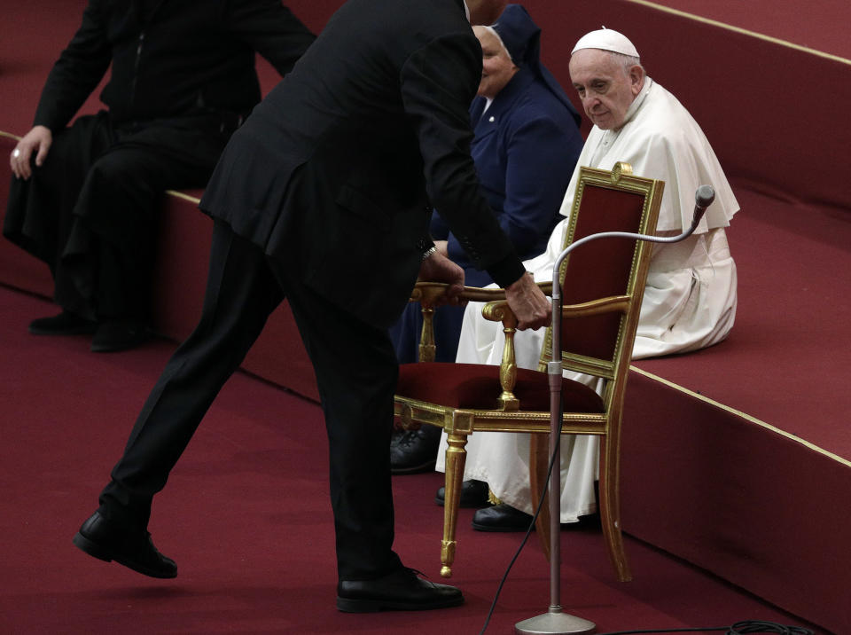 Pope Francis' butler Sandro Mariotti,left, removes a chair after the pontiff decided to sit on a step of a stage during audience with children and family from the dispensary of Santa Marta, a Vatican charity that offers special help to mothers and children in need, in the Paul VI hall at the Vatican, Sunday, Dec. 16, 2018. (AP Photo/Gregorio Borgia)