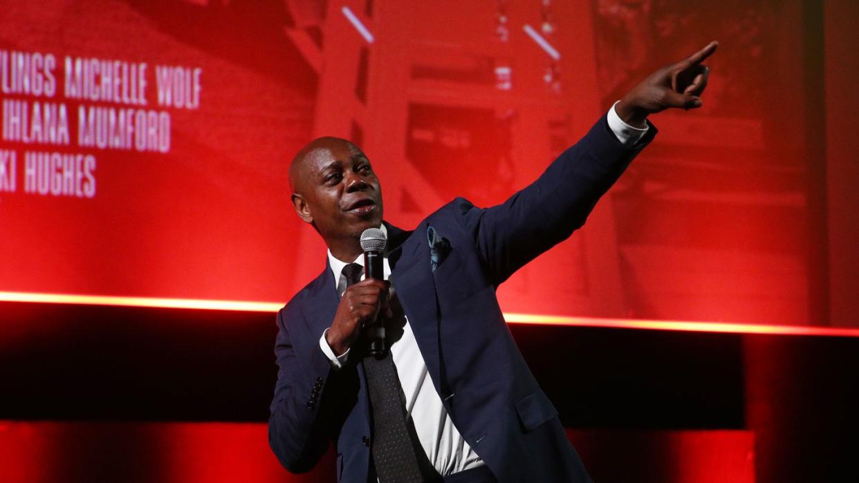 dave chappelle pointing upward with his left arm and hand and holding a microphone with the other