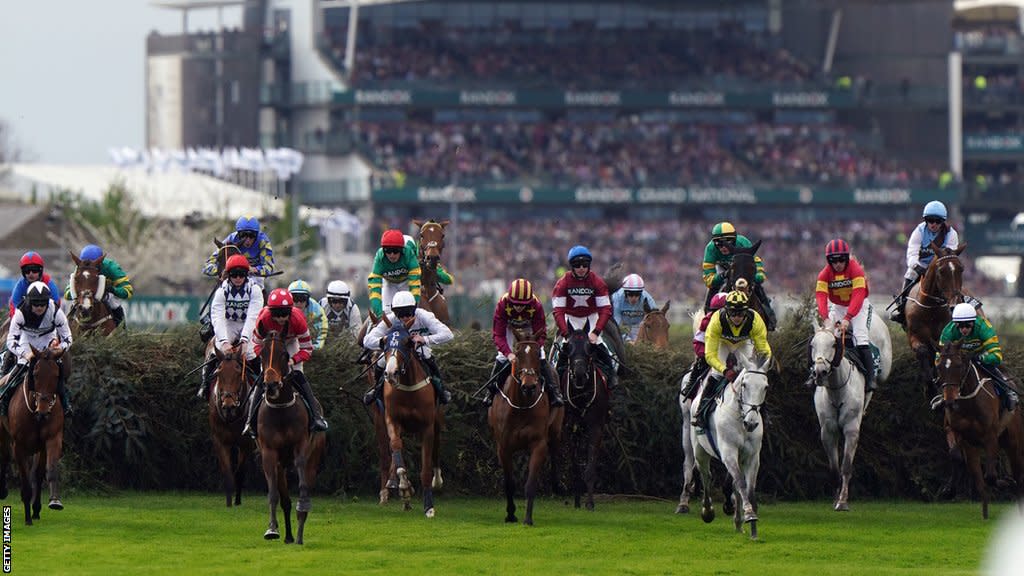 Horses jump a fence during the 2024 Grand National with the grandstands in the background