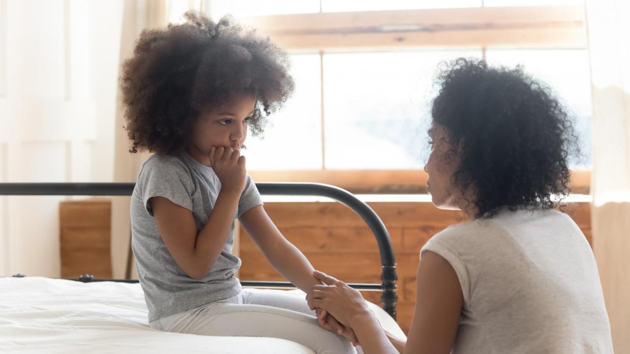 Caring worried african american mother holding hand of sad little mixed race daughter talking giving support and comfort, black mom foster parent consoling small kid being bullied sit on bed at home.