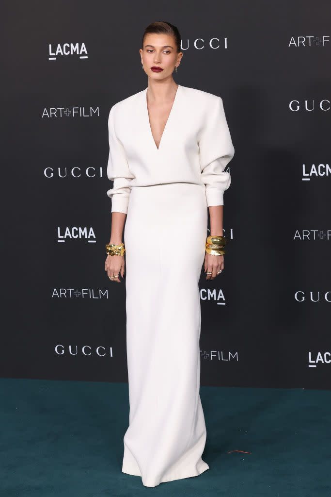 <p>Hailey Bieber gave us a lesson in pared-back red carpet glamour in a simple white column dress, featuring shoulder pads and a deep v-neck, by Saint Laurent to the LACMA Art + Film Gala. The 24-year-old paired the maxi dress with gold bangles and wore a deep red lip. </p>