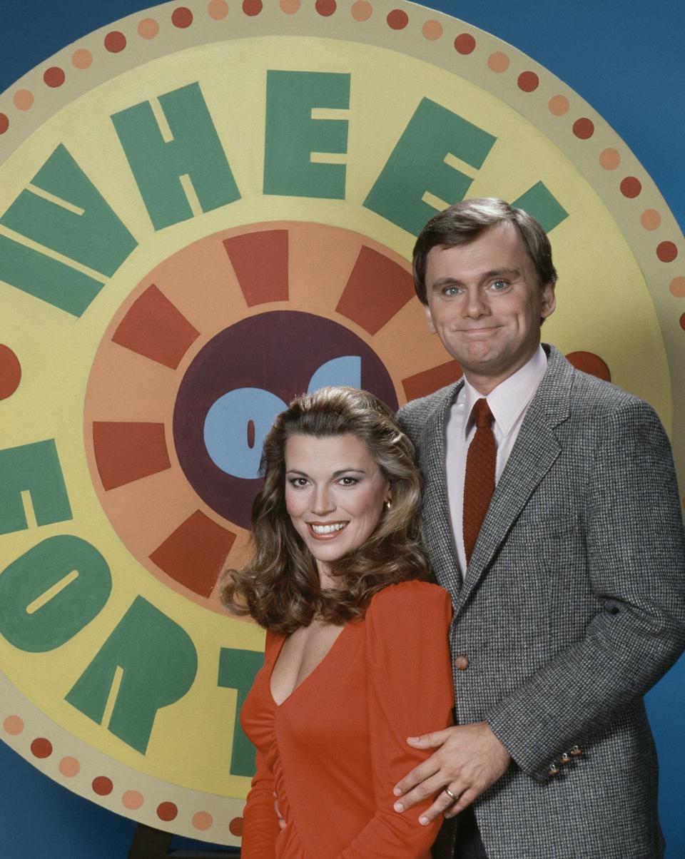 Vanna White wearing a red top, Pat Sajak holding her arm waring a suit with Wheel of Fortune logo behind them