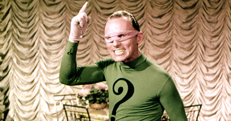 A still from Batman '66 shows Frank Gorshin as the Riddler wearing a pink domino mask and green suit with a large black question mark on it raising his hand menicingly