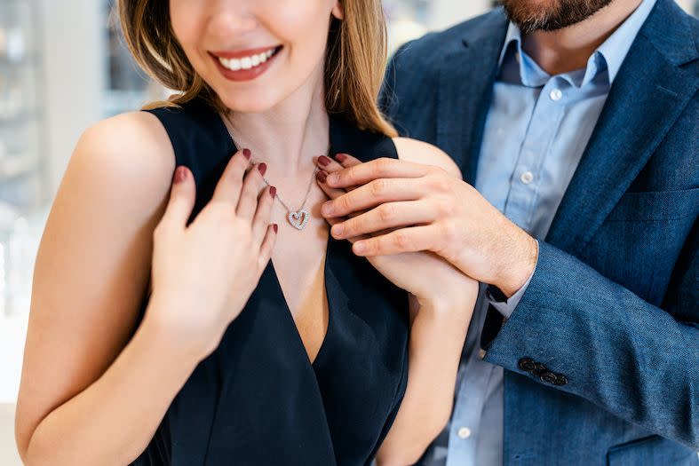 a young woman tries on a necklace as a man stands behind her and touches her hand which is placed over her chest